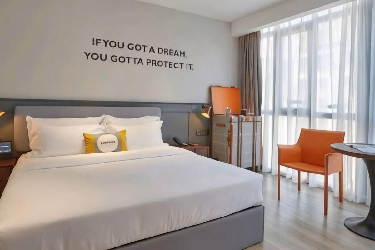 Habitación de hotel con cama, escritorio, silla, ventana con cortinas y una cita inspiradora en la pared.