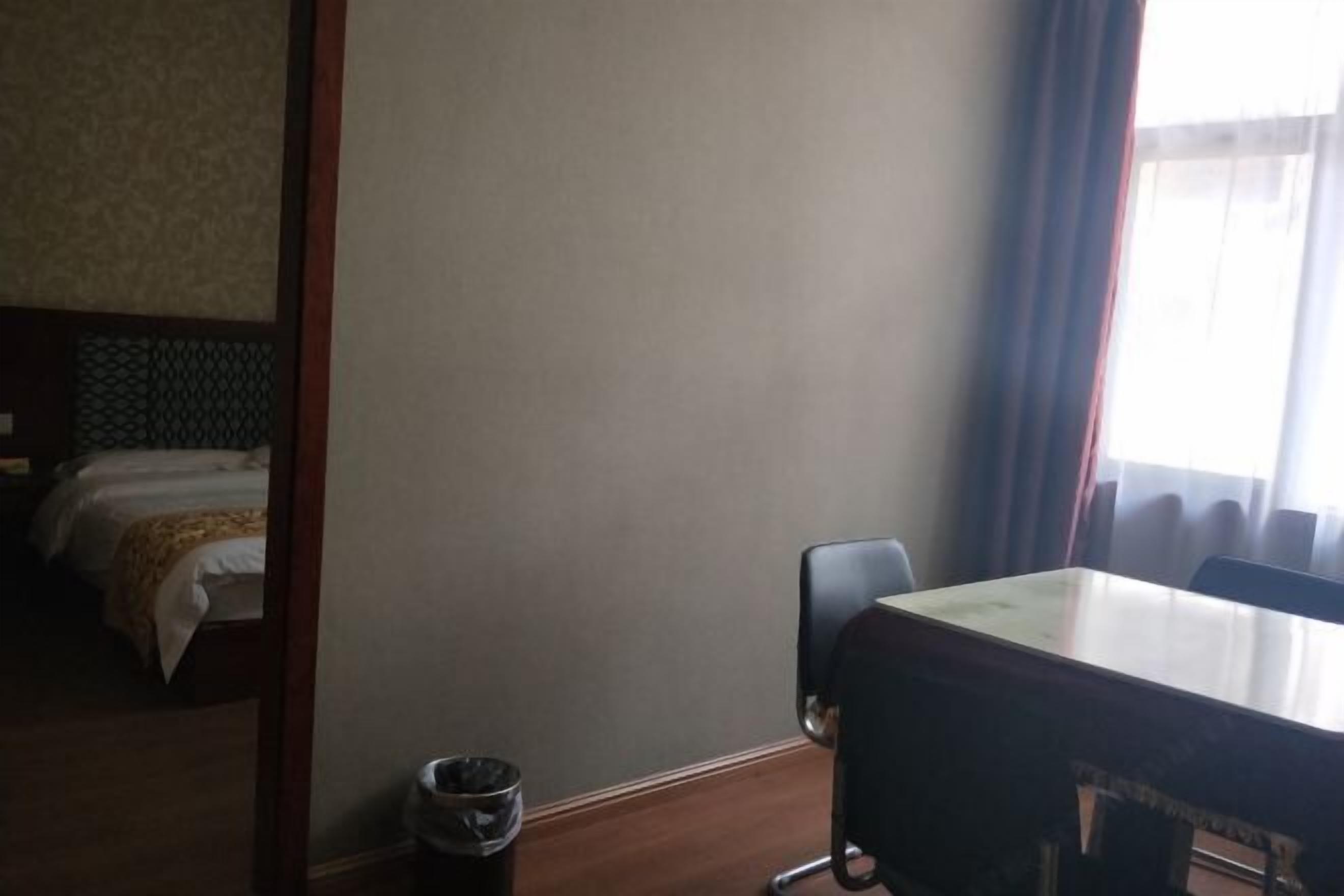 A hotel room with a bed, a wooden table, a chair, and a trash bin.