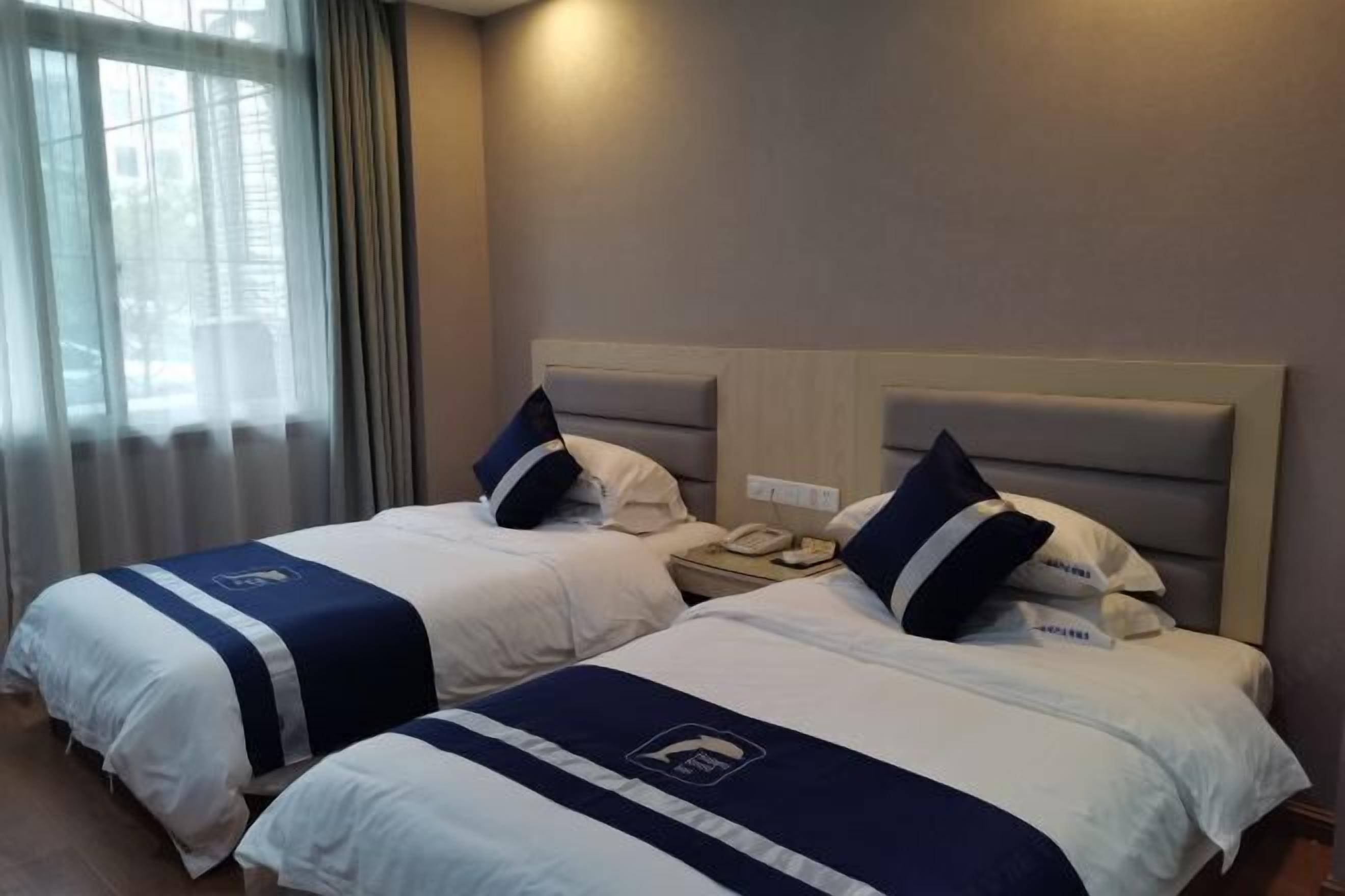 Habitación de hotel con dos camas, cada una con cubrecama y almohadas azules y blancas. Se observa una mesita de noche con un teléfono y una lámpara fijada a la pared.