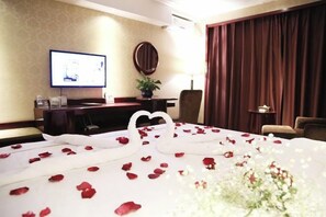 Une chambre d’hôtel avec un lit recouvert de pétales de roses, une télévision, un bureau avec un ordinateur et une grande fenêtre avec des rideaux.