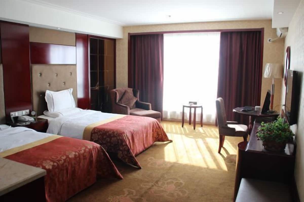 Une chambre d’hôtel avec deux lits, un bureau, une chaise, une petite table et une grande fenêtre avec des rideaux.