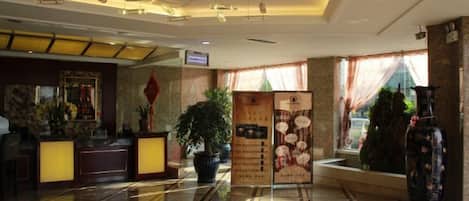 A hotel lobby with a reception desk, a large potted plant, a decorative vase, and a tiled floor.
