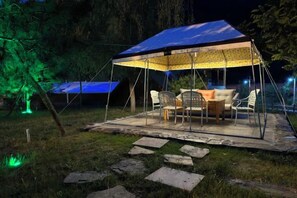 Ein Sitzbereich im Freien mit Zeltabdeckung, einem Tisch und Stühlen, umgeben von Steinstufen und Grünpflanzen.