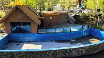 Una pequeña construcción con techo de paja y una ventana, junto a una piscina azul con borde de piedra.