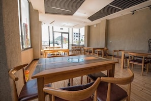 A dining area with wooden tables and chairs, large windows, and a view of buildings outside.