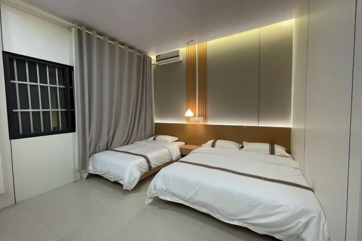 A hotel room with two single beds, a window with curtains, an air conditioning unit, and a bedside lamp.