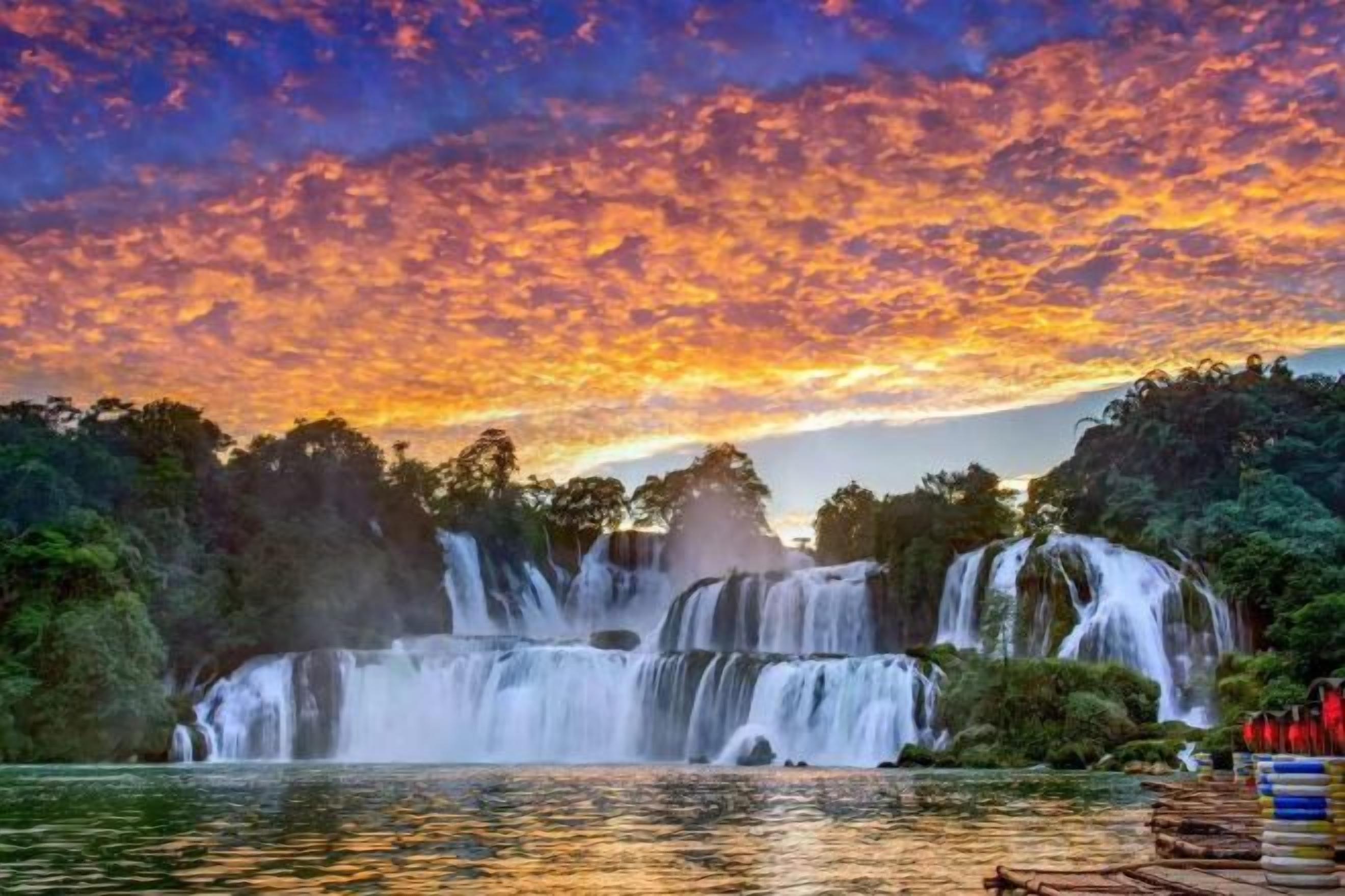 A waterfall with multiple tiers cascading into a body of water, surrounded by dense foliage and a vibrant sky.