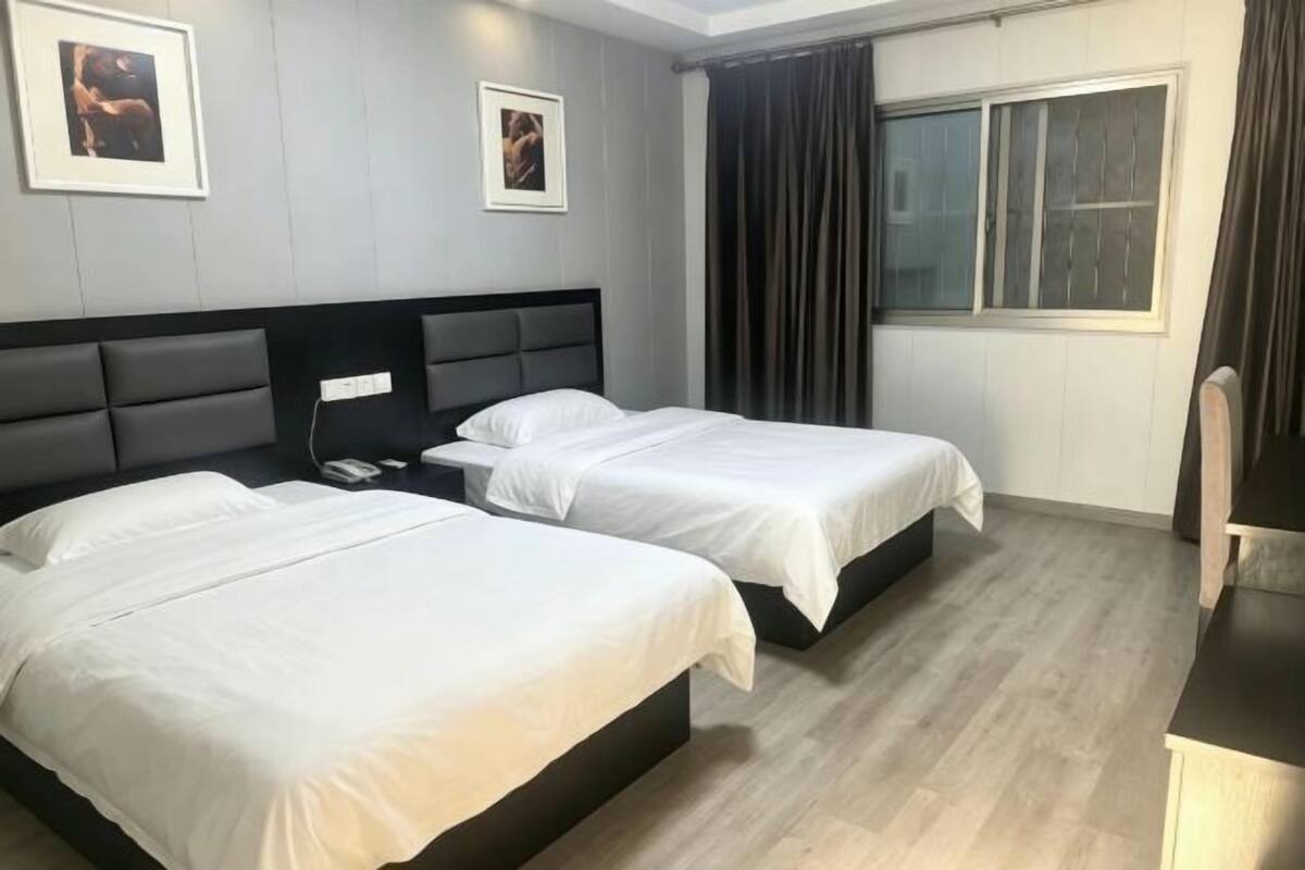 A hotel room with two beds, a desk, a chair, a window with curtains, and framed pictures on the wall.