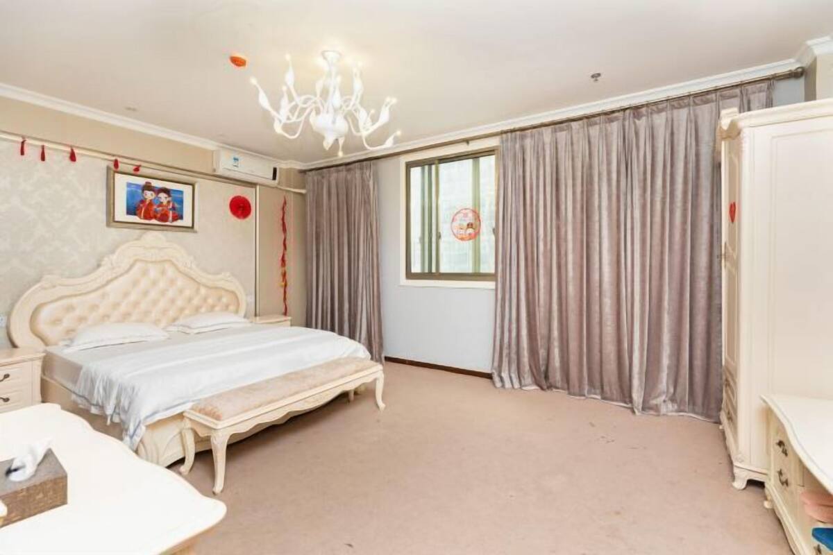 A bedroom with a large bed, a bedside table, a dresser, a chandelier, and curtains.