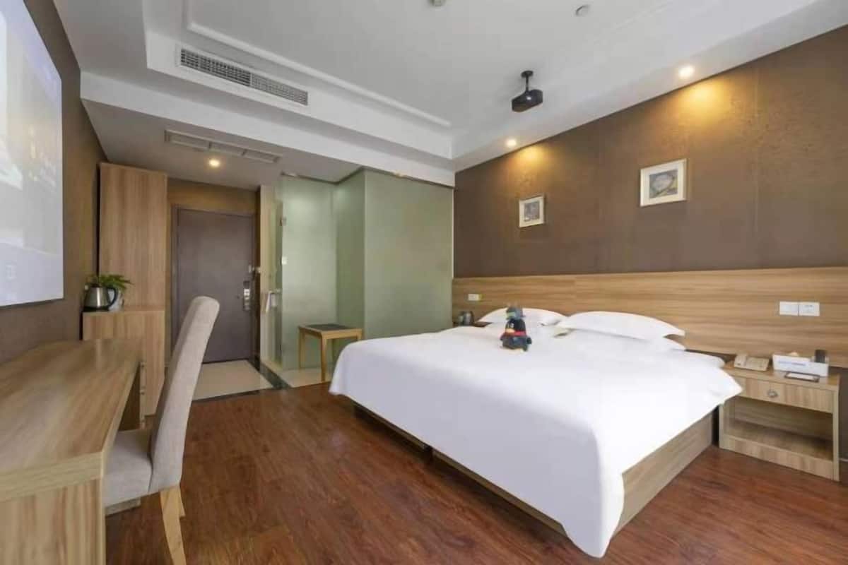 A hotel room with a wooden floor, a bed with white bedding, a desk with a chair, and a small bedside table with a lamp.