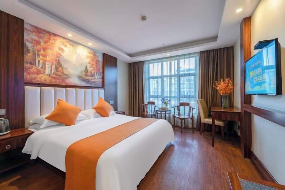 A hotel room with a large bed, a desk, and a chair. There is a painting on the wall and a TV on the wall.