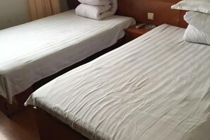A hotel room with two single beds, each with a white bedspread and pillows. There is a wooden headboard on the right side of the bed, and a small wooden nightstand with a glass on it.