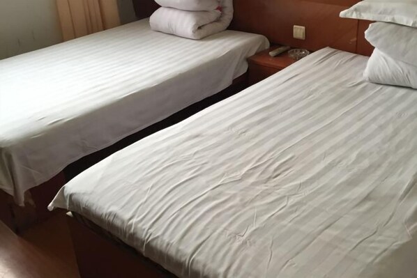 Una habitación de hotel con dos camas individuales, cada una con una colcha y almohadas blancas. Se observa un cabecero de madera en el lado derecho de la cama, y una mesita de noche de madera con un vaso encima.