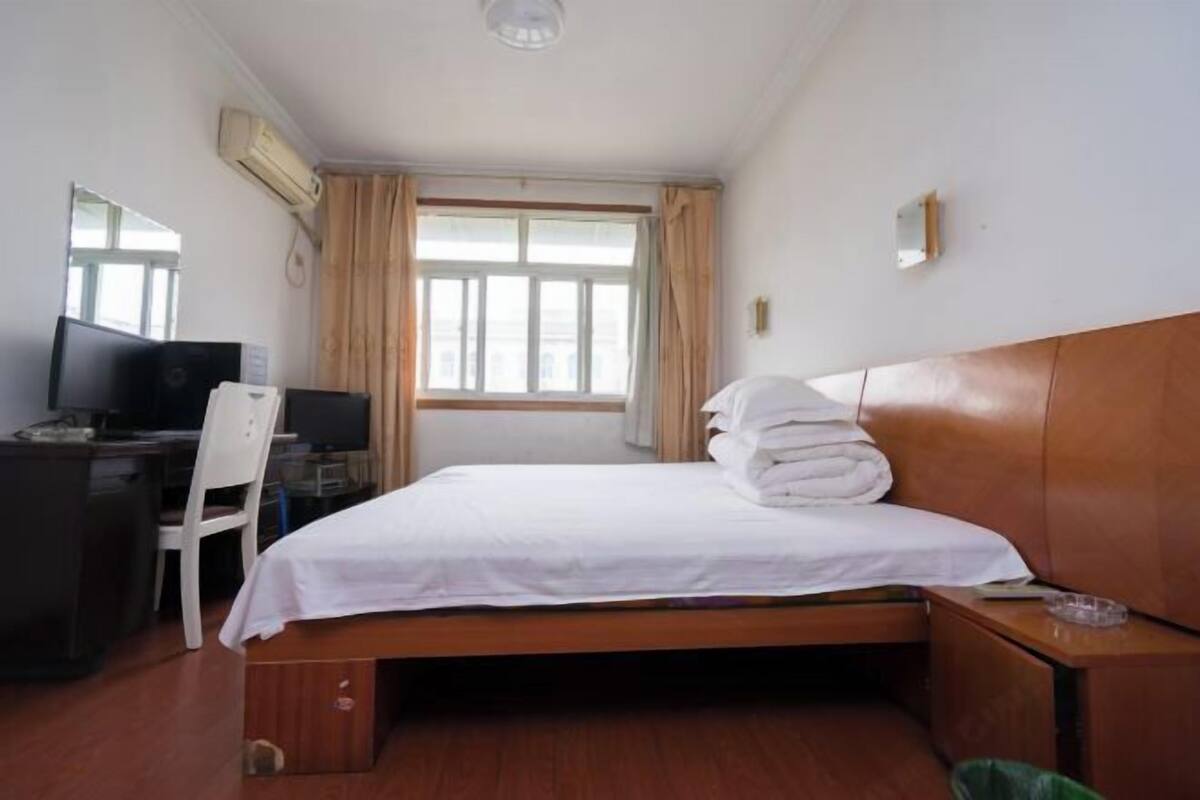 Hotellihuone, jossa on sänky, työpöytä tietokoneella, tuoli, ikkuna verhoilla ja ilmastointilaite.