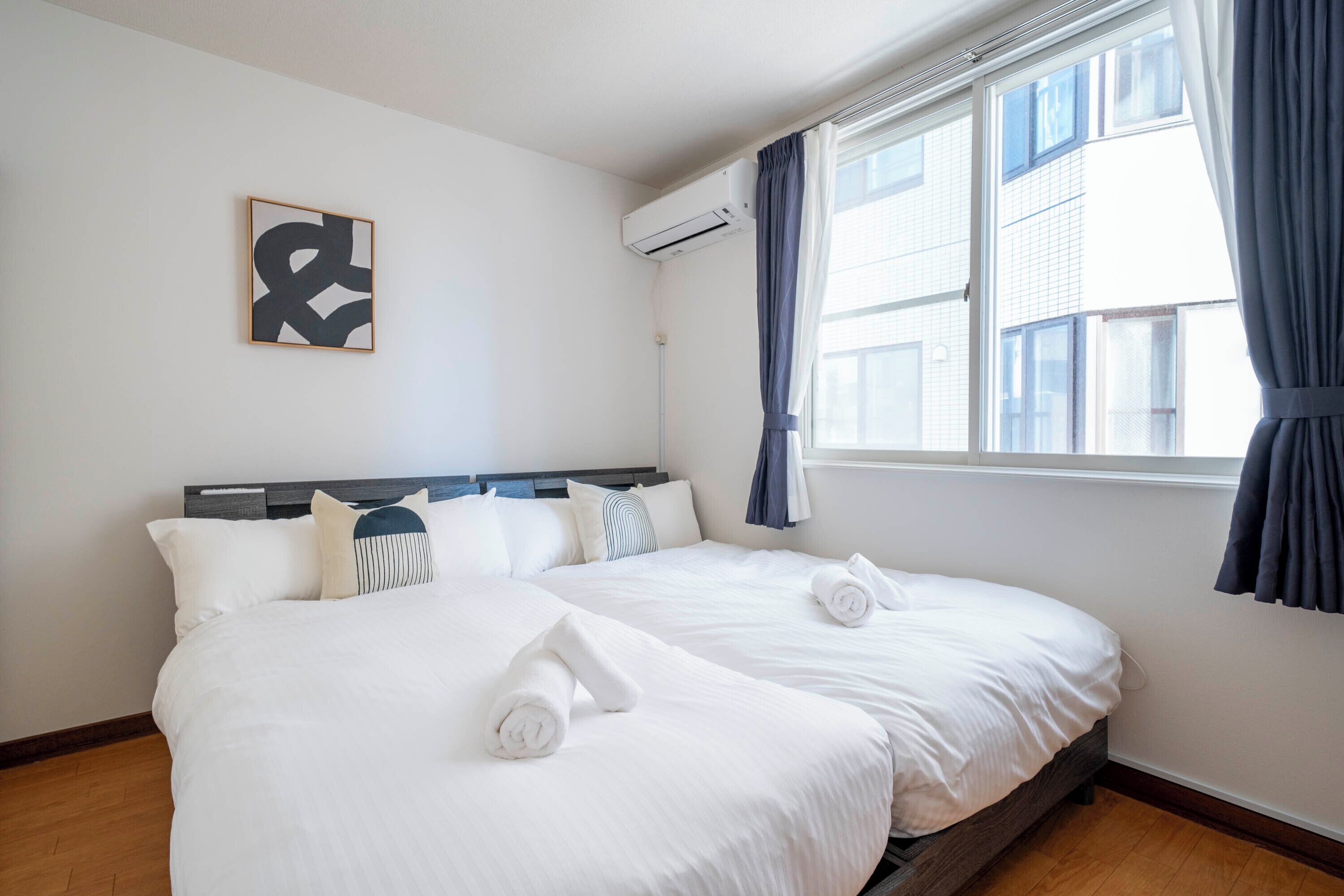 A hotel room with two beds, a large window with curtains, an air conditioning unit, and a framed abstract artwork on the wall.