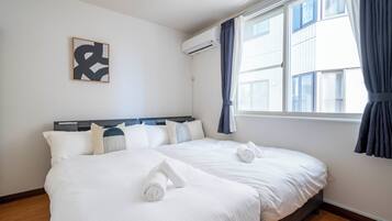 A hotel room with two beds, a large window with curtains, an air conditioning unit, and a framed abstract artwork on the wall.