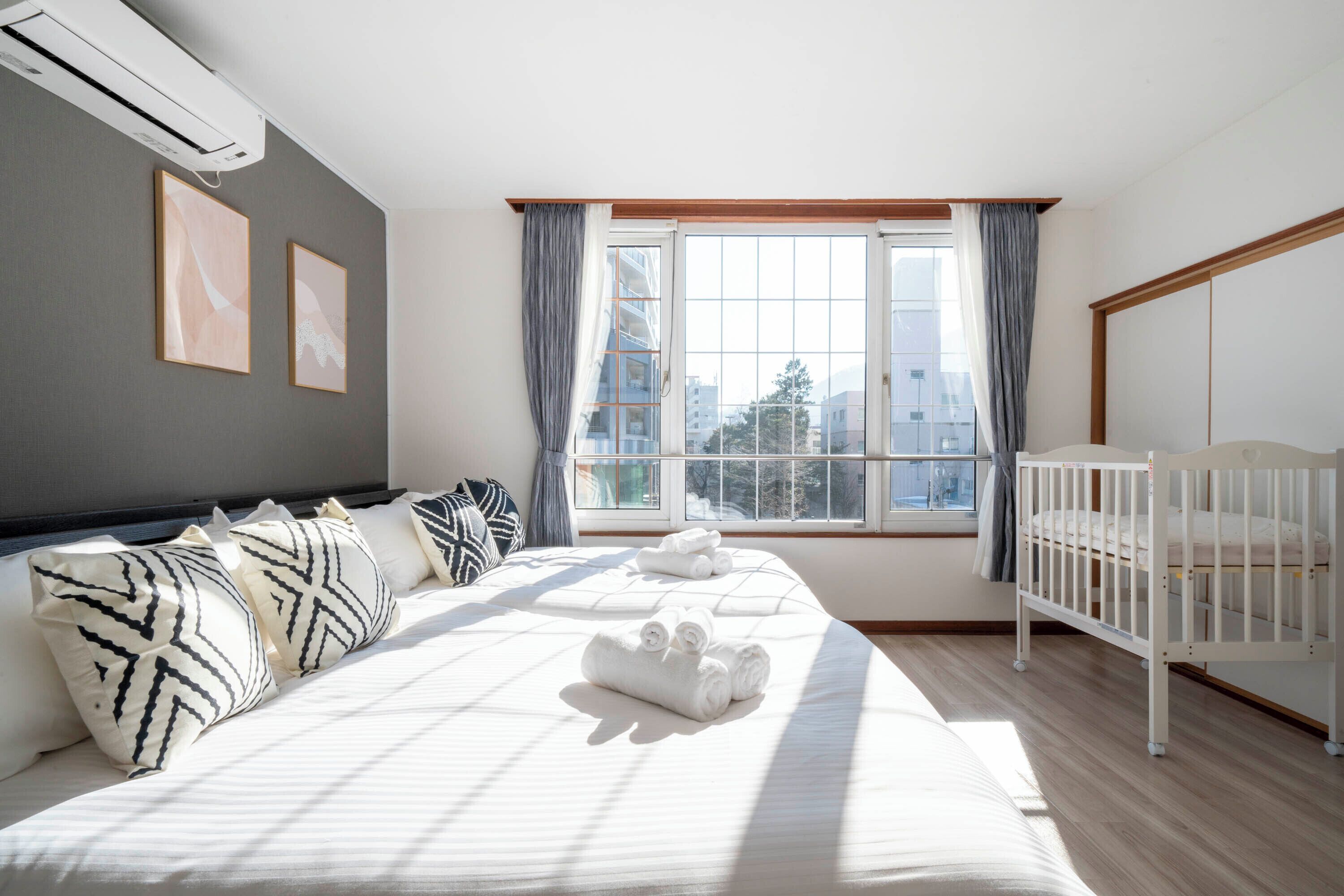 Une chambre à coucher avec un lit, des oreillers, un berceau, une fenêtre et un appareil de climatisation.