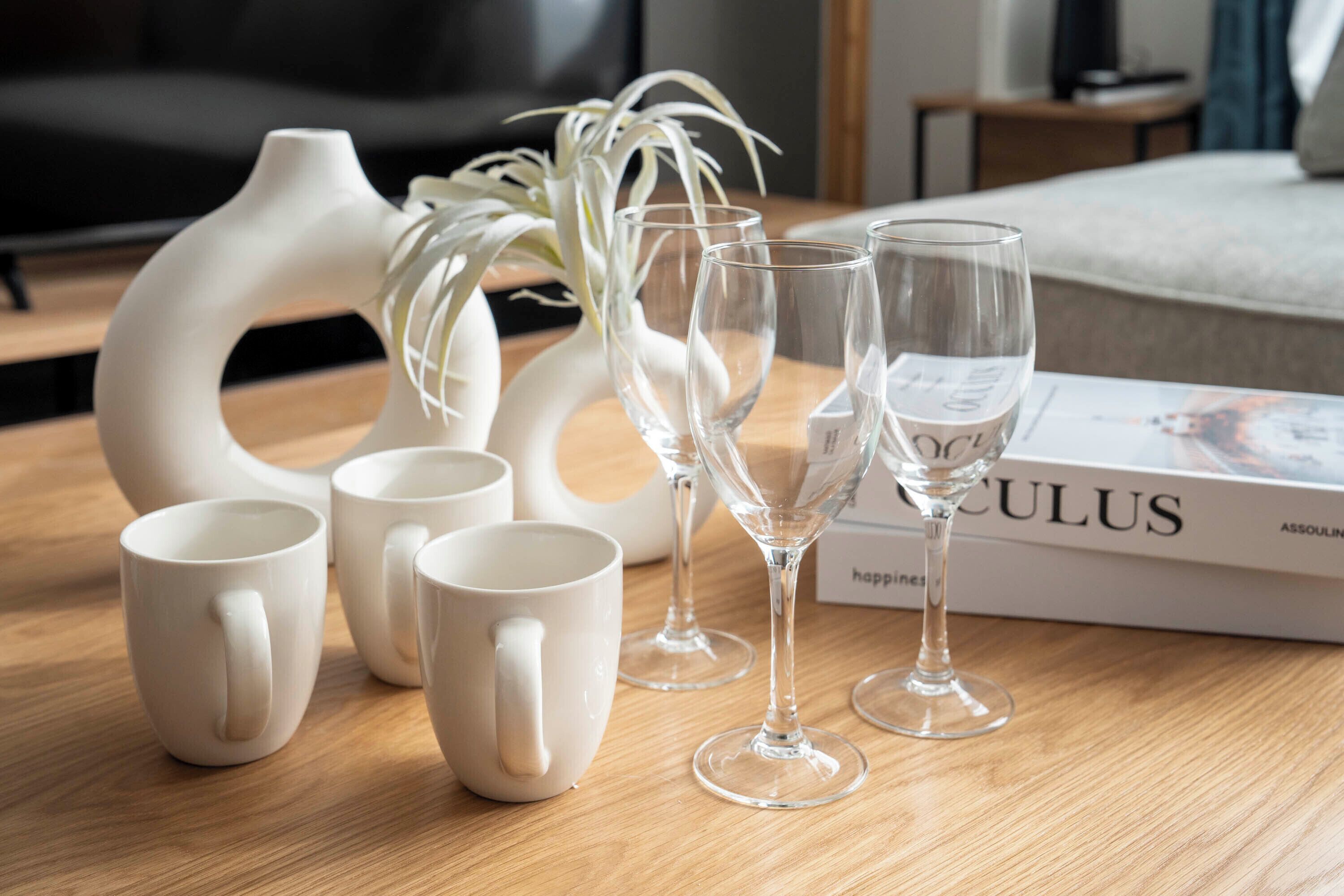 A wooden table with white ceramic mugs, clear wine glasses, and a white vase with a plant. A book titled 'Calculus' is visible in the background.