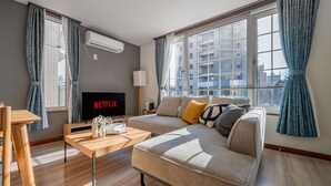 Ein modernes Wohnzimmer mit einem Ecksofa, einem Holztisch, einem Flachbildfernseher, der 'NETFLIX' anzeigt, und großen Fenstern mit Vorhängen.