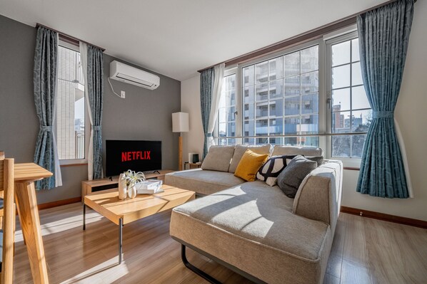 A modern living room with a sectional sofa, a wooden coffee table, a flat-screen TV displaying 'NETFLIX', and large windows with curtains. - GRAND SOLEIL (Sapporo)