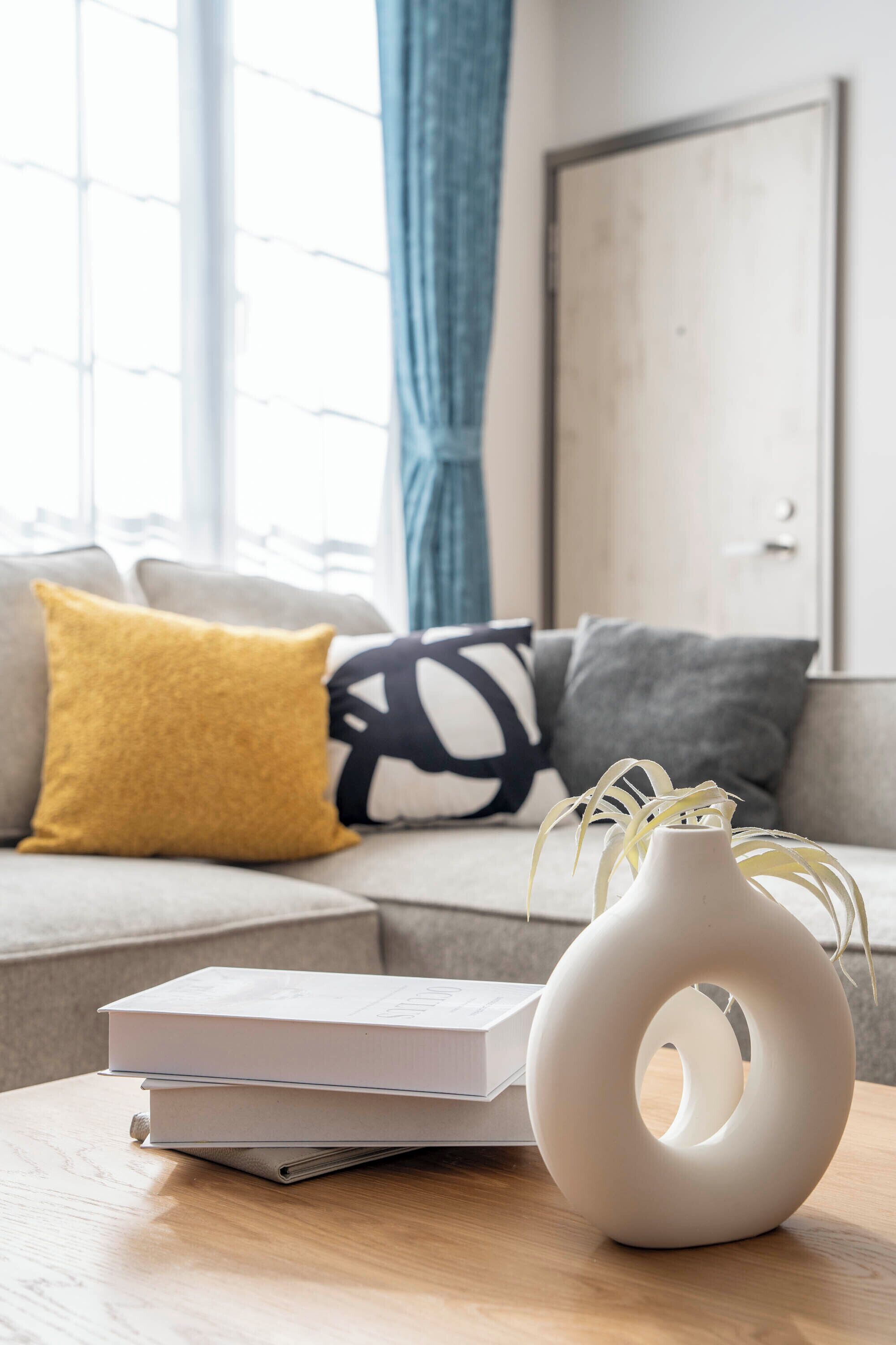 A modern living room with a sofa, cushions, a white vase, and books on a wooden table.