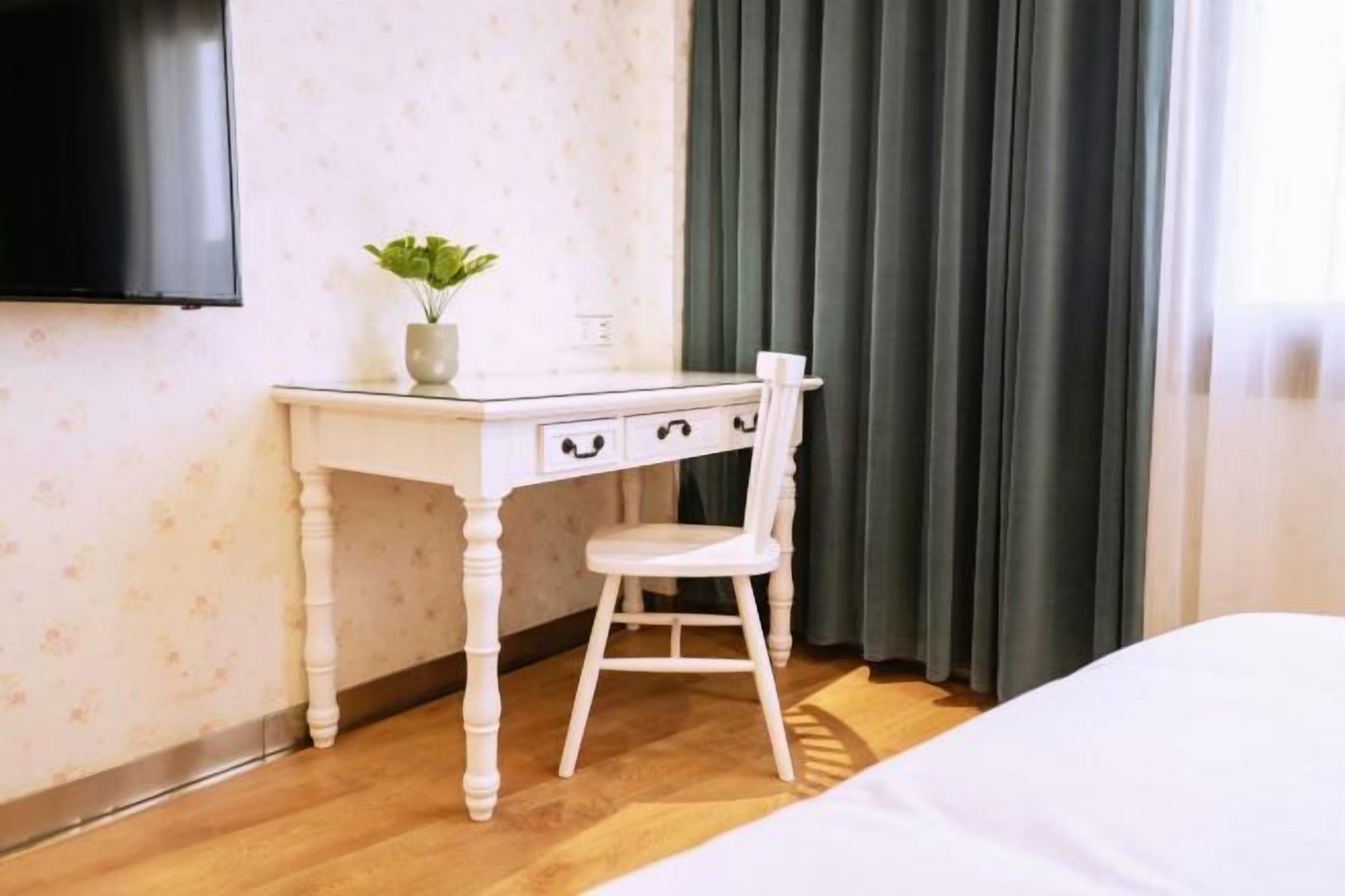 Quarto com cama, escrivaninha com cadeira, planta em vaso e televisão na parede.