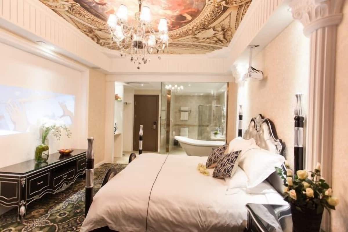 A spacious bedroom with a large bed, a chandelier, a glass-enclosed bathroom, and a decorative ceiling.