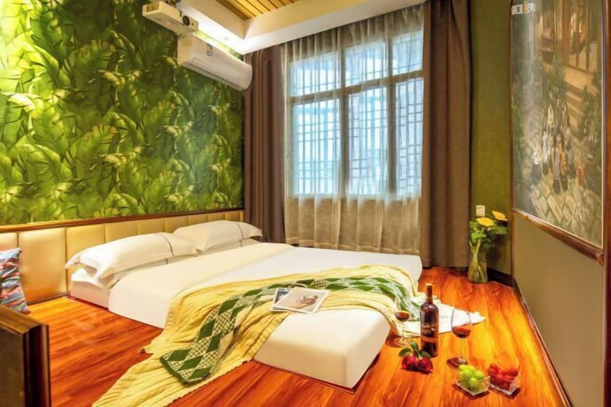 A hotel room with a bed, a wooden floor, a green wall, a window with curtains, and a painting on the wall.