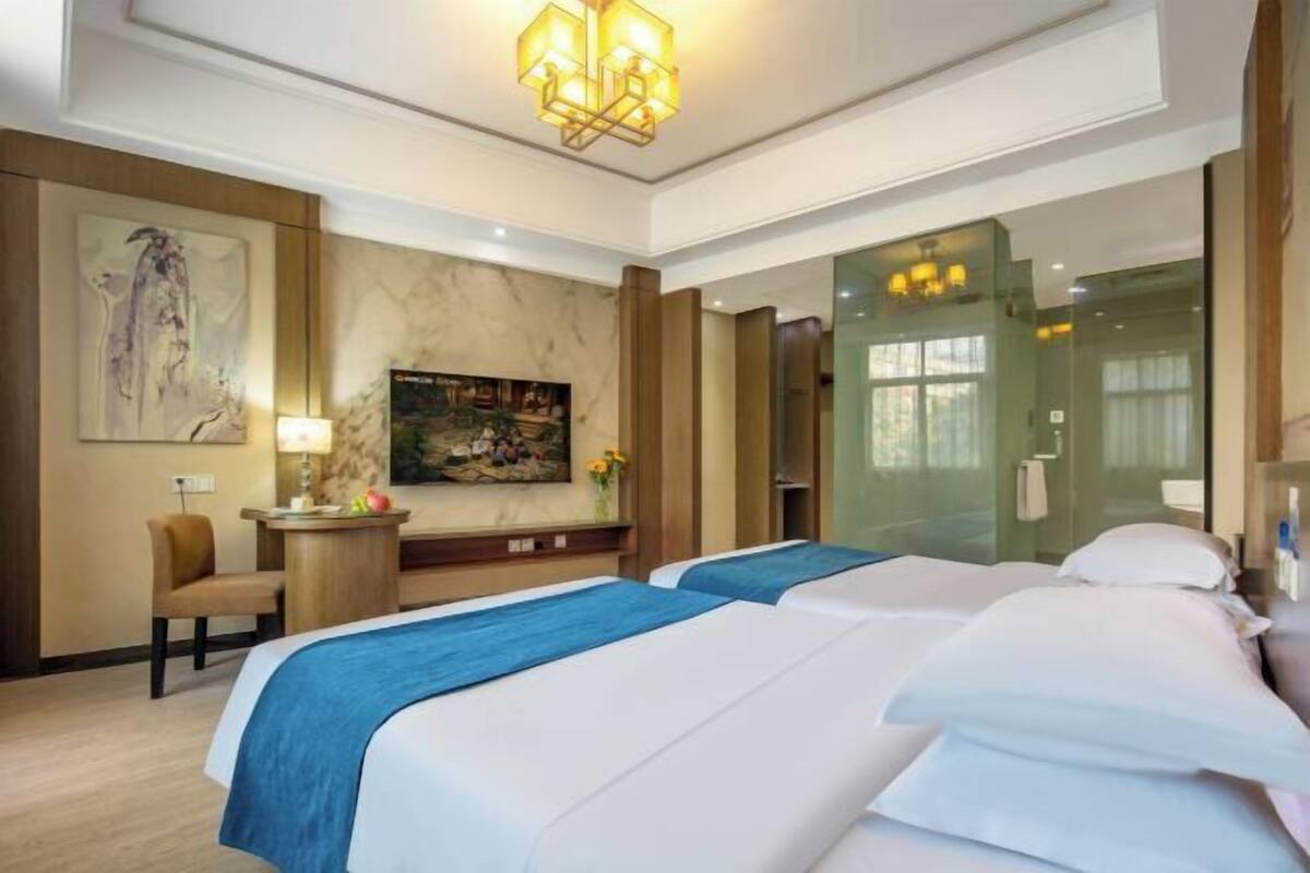 A hotel room with two beds, a desk, a chair, a painting, and a glass-enclosed shower area.