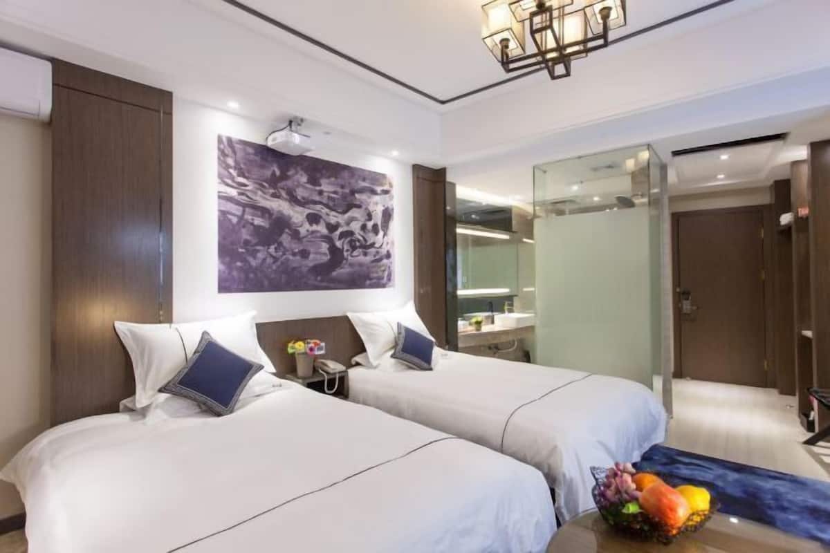 A hotel room with two beds, a large abstract painting, a glass shower enclosure, and a fruit bowl on the side table.