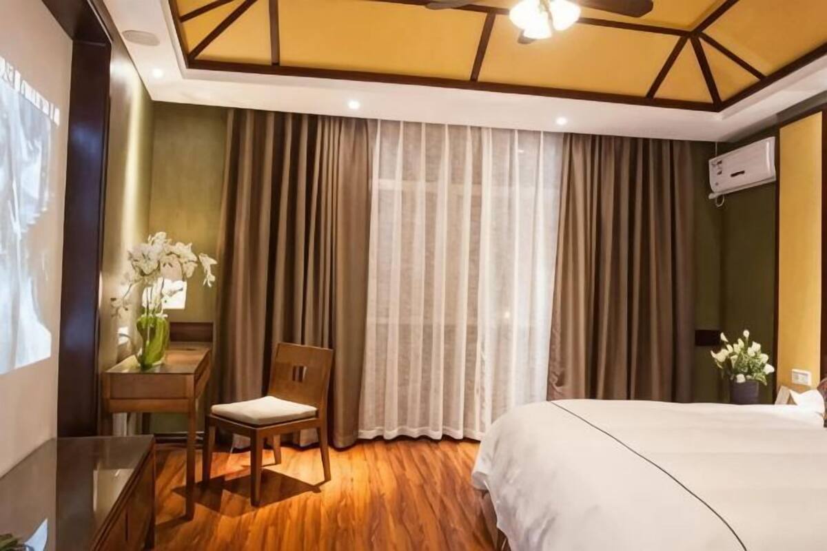A hotel room with a bed, a desk with a chair, a vase with flowers, and a large window with curtains.