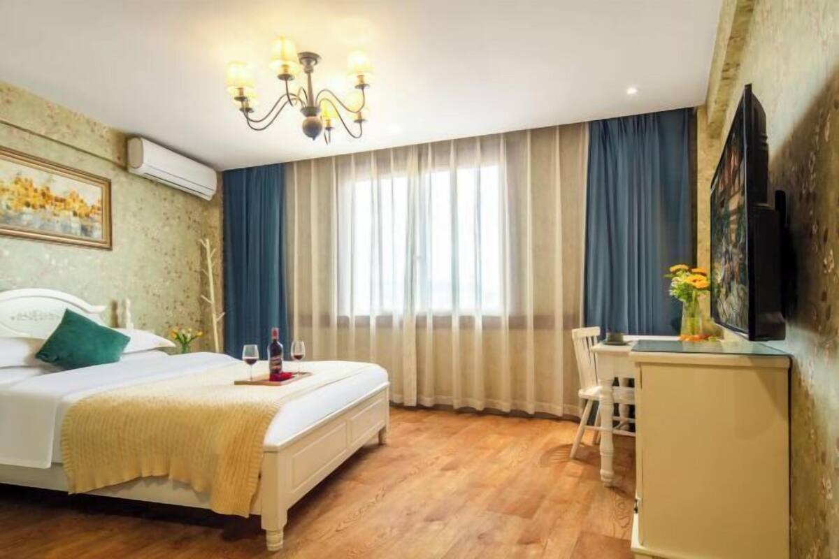 A hotel room with a bed, a desk, a chair, a chandelier, and a large window with curtains.