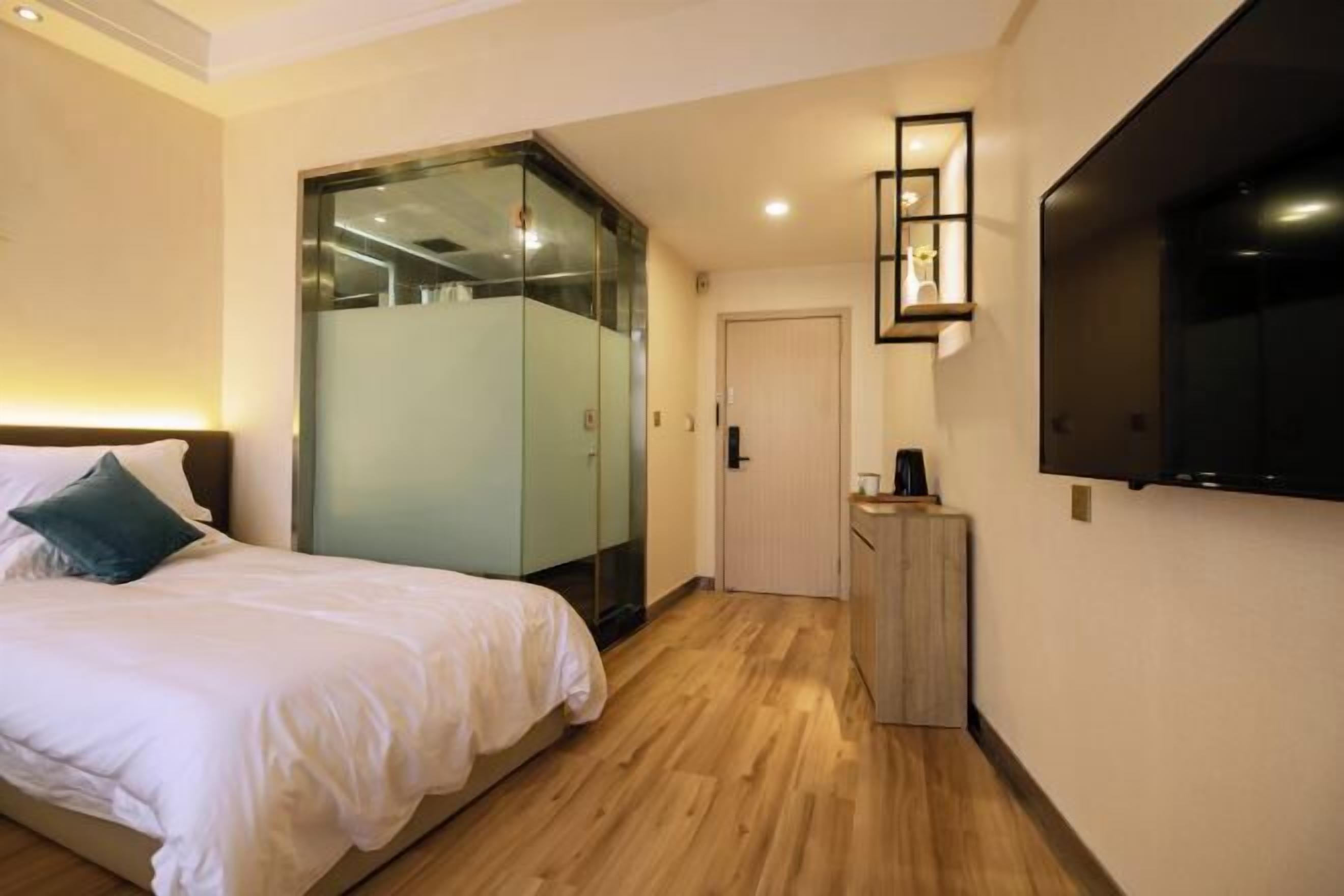 A hotel room with a bed, a glass wardrobe, a small wooden cabinet, and a TV mounted on the wall.
