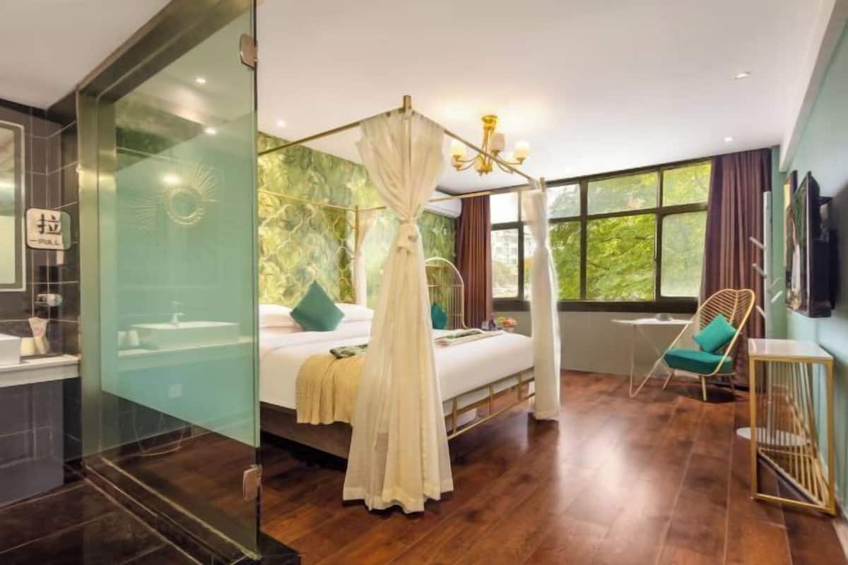 A bedroom with a canopy bed, a sitting area with a chair, a TV, and a large window.