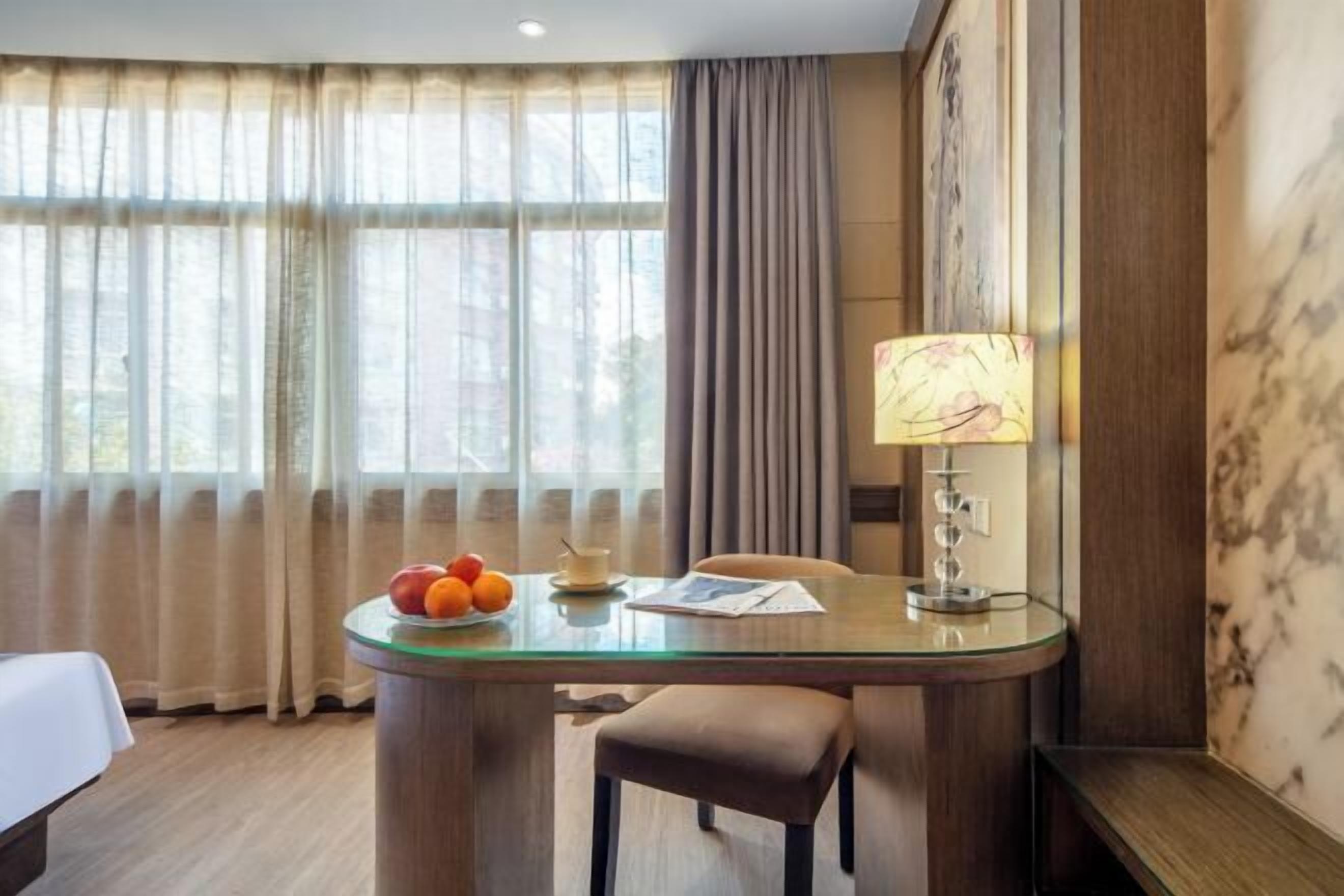 Ein Hotelzimmer mit Glastisch, Lampe, Stuhl, Bett und einem Fenster mit Vorhängen.