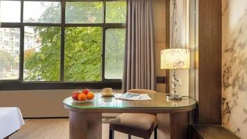 A hotel room with a glass-top table, a lamp, a chair, and a view of trees through large windows.