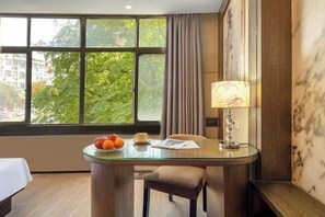 Habitación de hotel con una mesita de vidrio, una lámpara, una silla y vistas a árboles a través de amplias ventanas.