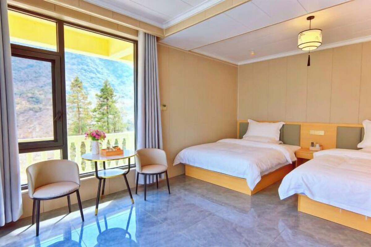 Ein Hotelzimmer mit zwei Betten, einem kleinen Tisch, zwei Stühlen, einem Fenster mit Blick auf Bäume und einer Deckenleuchte.