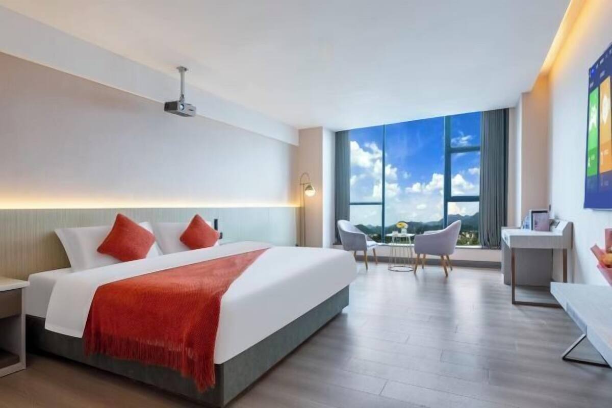A modern hotel room with a large bed, a desk, and a view of the sky through a large window.