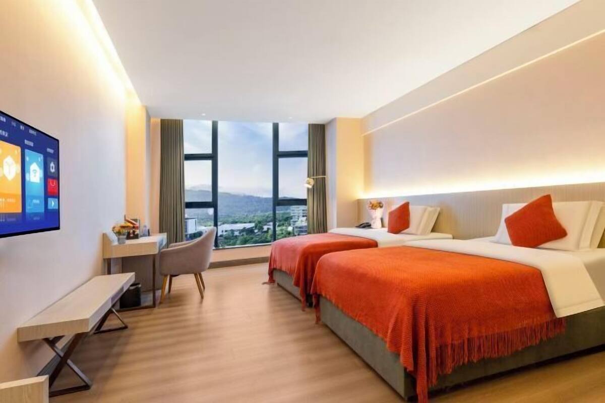 A hotel room with two beds, a desk, a chair, a TV, and a large window with a city view.
