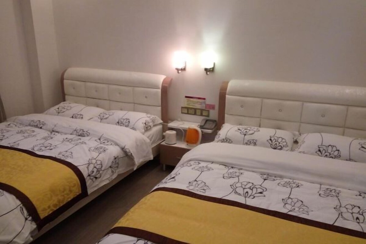A hotel room with two beds, each with floral-patterned bedding and a headboard. There is a nightstand with a lamp, a telephone, and a small table with a book and a cup.