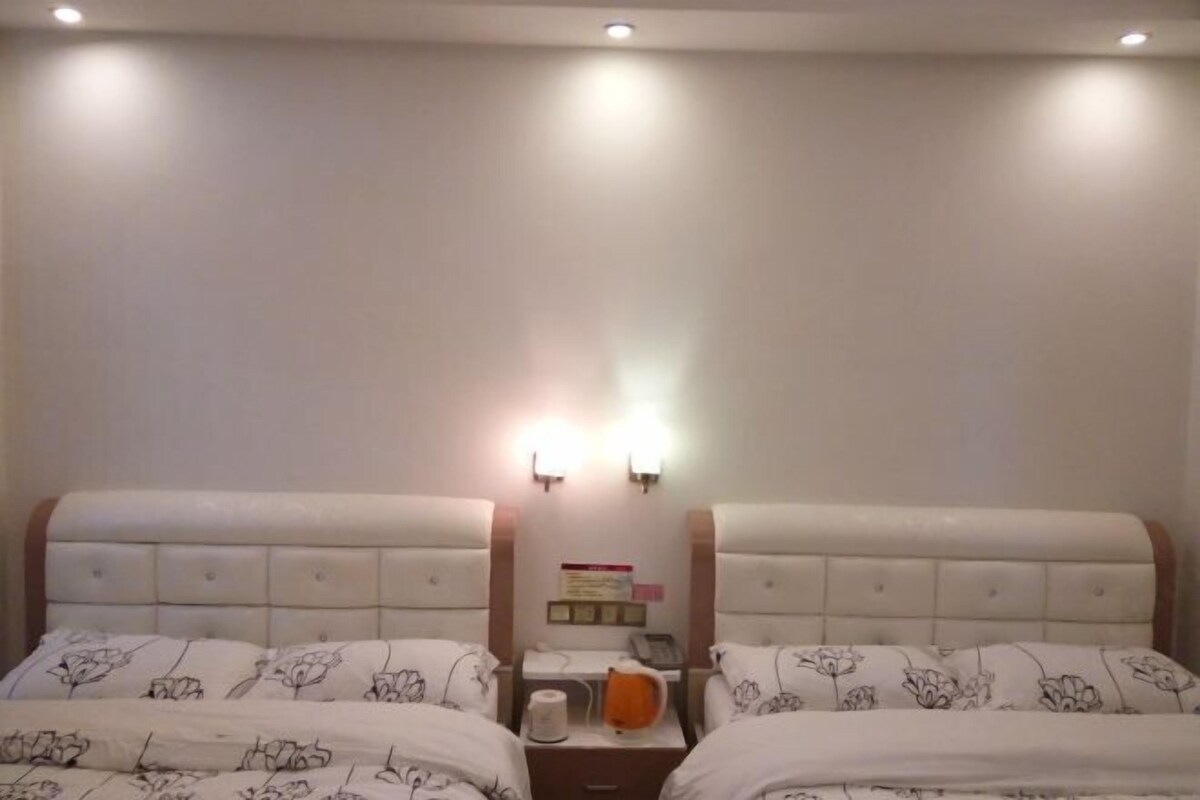 A hotel room with two beds, each with a floral-patterned duvet, a bedside table with a kettle and a phone, and a wall-mounted light fixture.