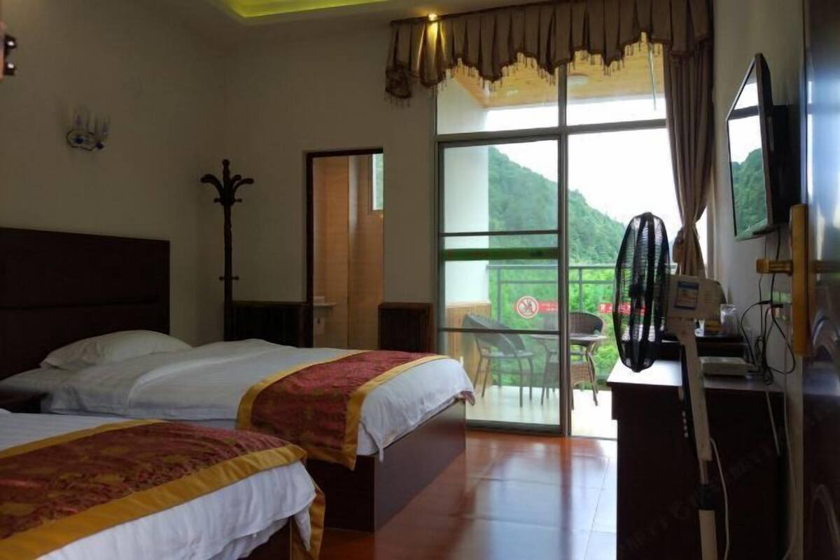Quarto de hotel com duas camas, uma janela grande com vista para o verde, uma mesa com ventilador e uma televisão.