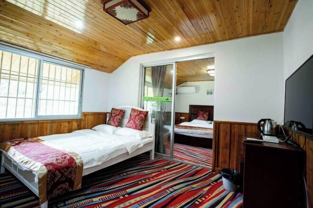 Quarto com teto de madeira, cama com colcha vermelha e branca, tapete colorido listrado e uma televisão sobre um suporte de madeira.