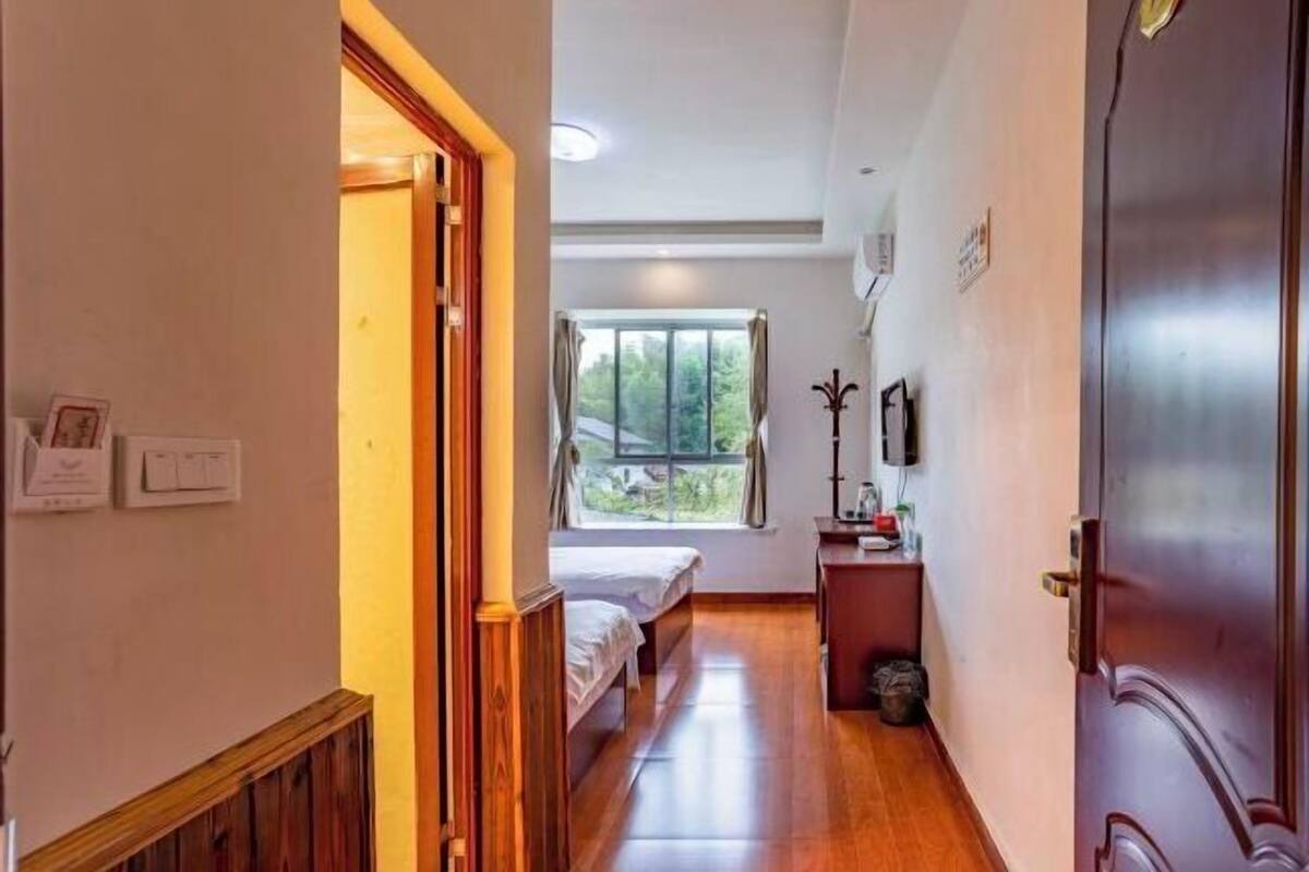 Um corredor estreito que leva a um quarto, onde se vê uma cama, uma janela e uma pequena mesa com abajur.