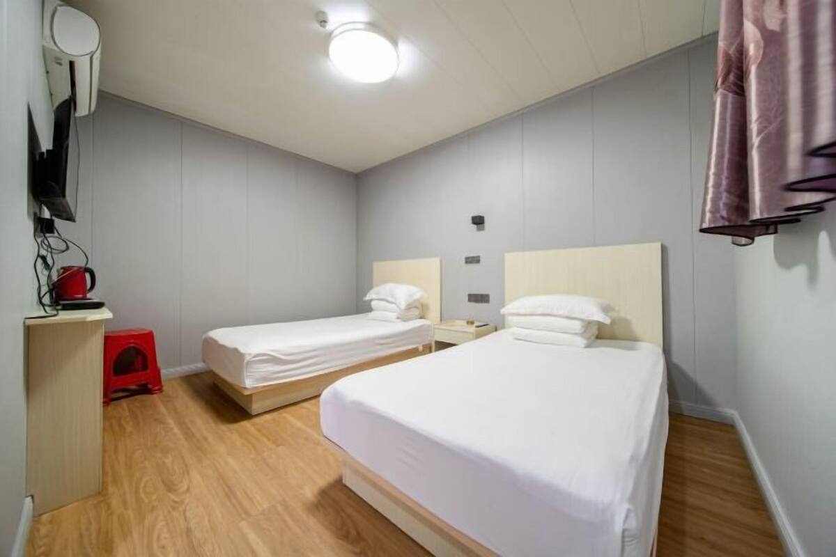 A hotel room with two single beds, a small desk with a red stool, a wall-mounted heater, and a ceiling light.