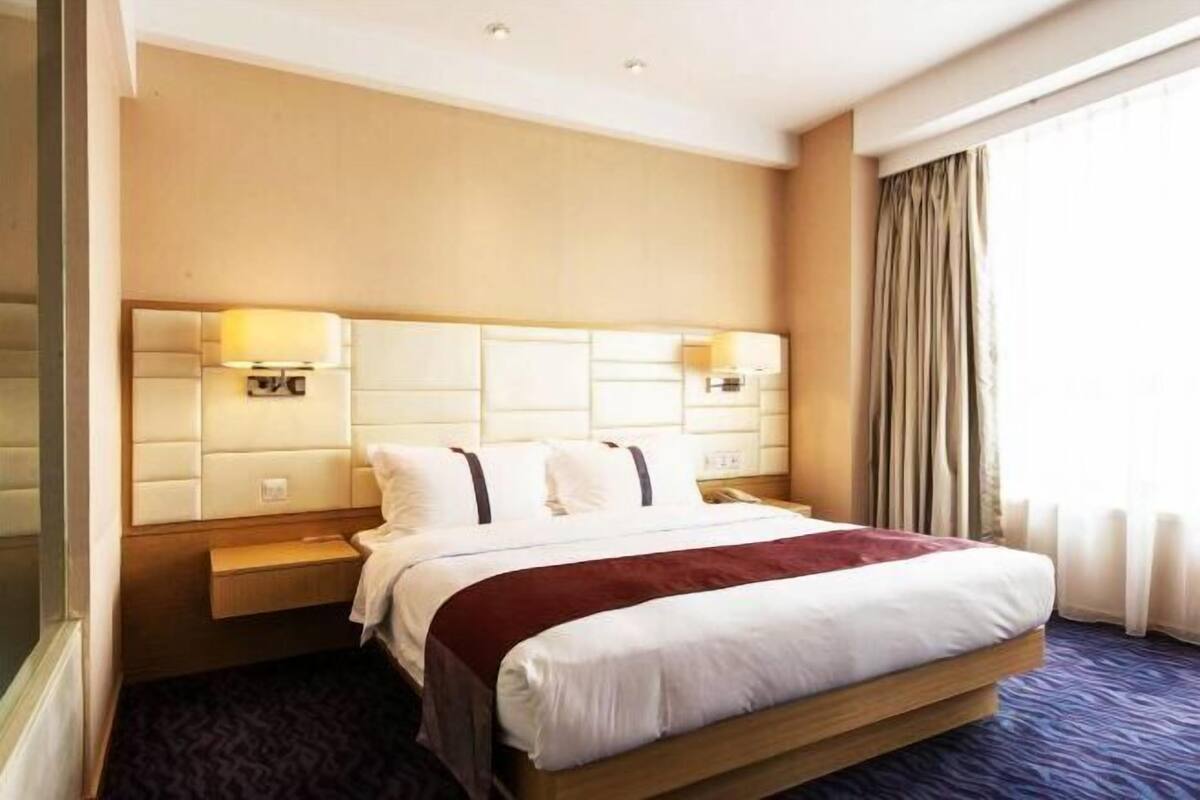 A hotel room with a large bed, headboard with built-in lighting, a bedside table, and a window with curtains.