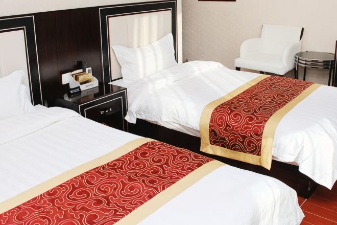 Kaksi sänkyä valkoisilla lakanoiden ja punaisilla sekä kultaisilla peitteillä, yöpöytä lampun kanssa ja valkoinen tuoli hotellihuoneessa.
