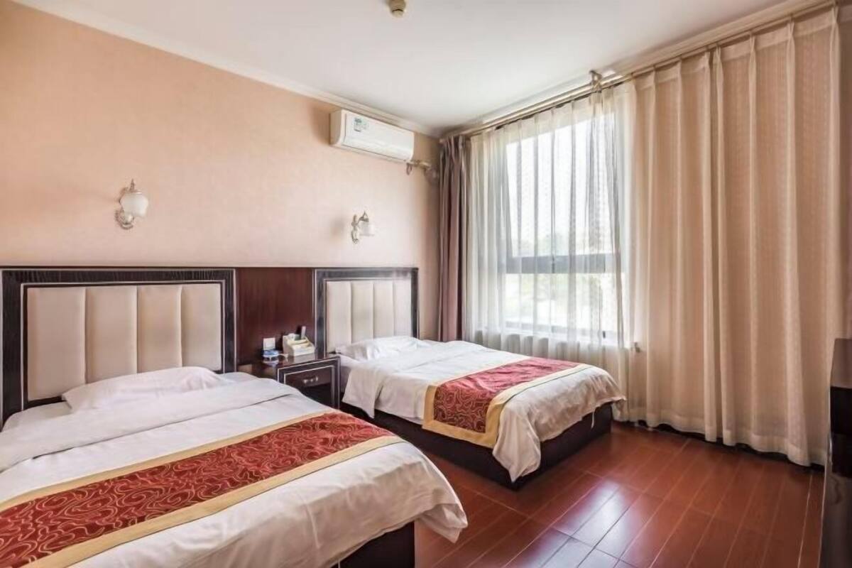 A hotel room with two beds, a wooden floor, a window with curtains, and an air conditioning unit.