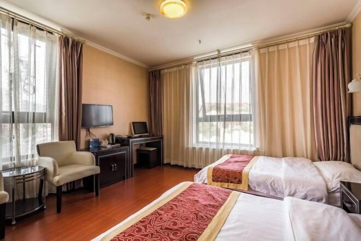 A hotel room with two beds, a desk, a chair, a TV, and large windows with curtains.
